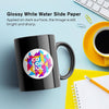 Glossy white waterslide decal paper applied to a black mug, showcasing a colorful design on a bright workspace.