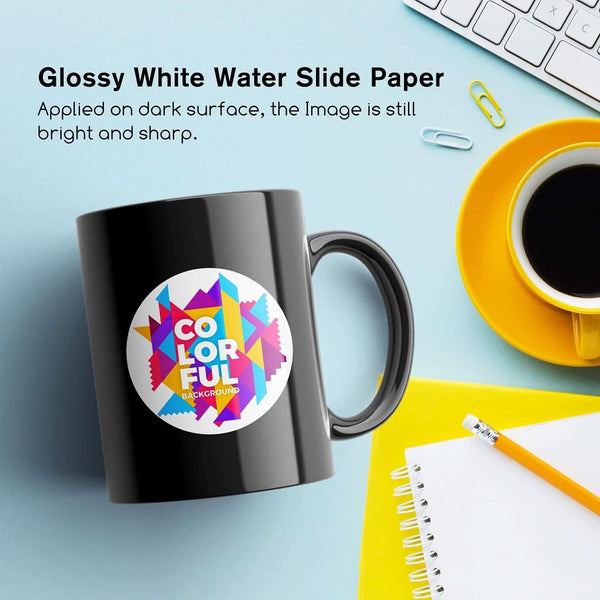 Glossy white waterslide decal paper applied to a black mug, showcasing a colorful design on a bright workspace.